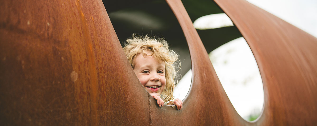 A little curly haired girl steals a glance out of a round hole in the side of rusty structure.